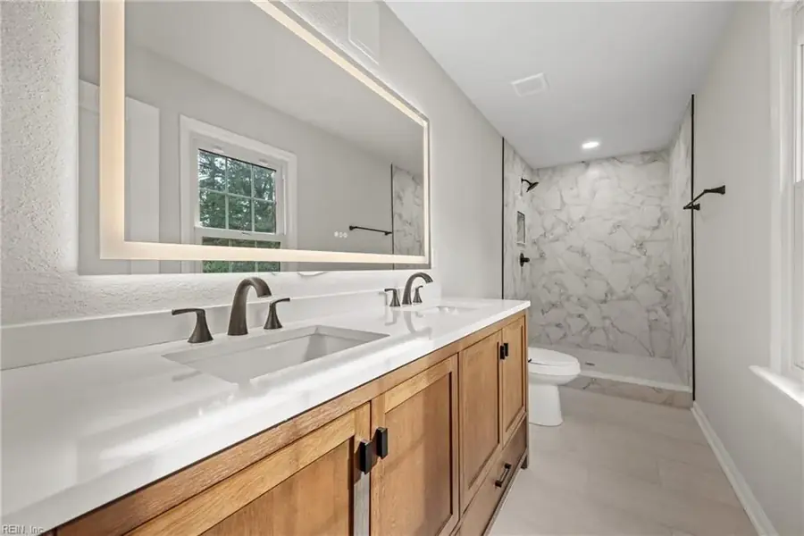 Bathroom with a wood double vanity, backlit mirror and marble-look shower surround